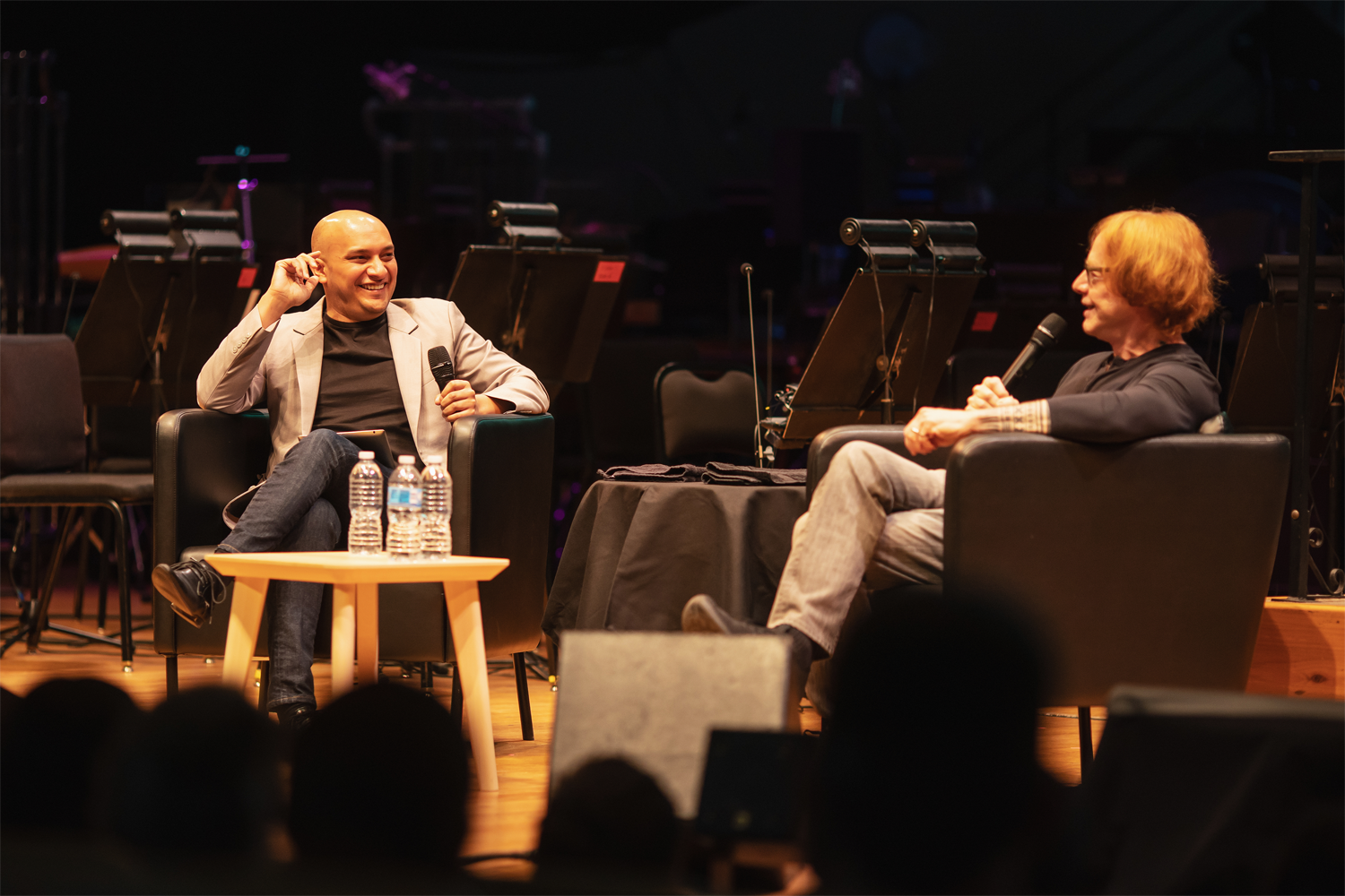 Christopher Dragon and Danny Elfman seated in black armchairs on the stage of Boettcher Concert Hall, speaking with microphones during a Prelude pre-concert talk in January 2022. A small wooden side table with water bottles sits between them. Empty orchestra chairs and music stands are arranged in the background. Photo credit: Amanda Tipton.