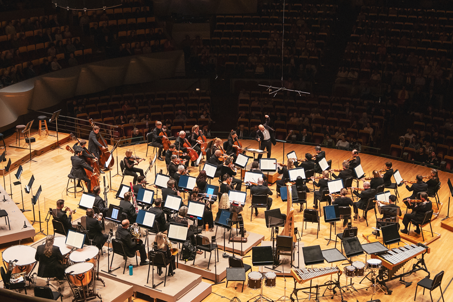 An overhead view of Christopher Dragon conducting a full orchestra during a Classics performance of Beethoven's Symphony No. 2 in 2025. Dragon stands at the podium near center stage with one arm raised, surrounded by musicians in black formal attire arranged in the traditional orchestral layout. Music stands with blue and white score folders fill the warm, wood-toned stage of Boettcher Concert Hall. Audience members are visible in the tiered seating surrounding the in-the-round stage. Photo credit: Amanda Tipton.