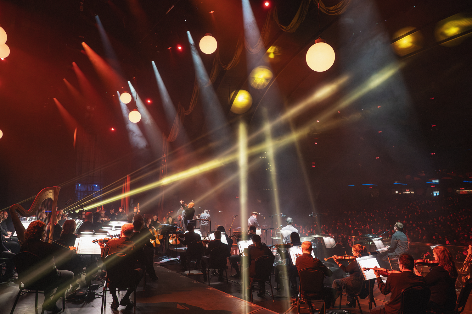 A wide-angle view from the side of the stage at Radio City Music Hall in January 2026, capturing Christopher Dragon conducting a sold-out performance with Gregory Alan Isakov. Dramatic beams of golden and white light cut across the darkened venue from above, illuminating the orchestra and its music stands below. A harp is visible on the left, with strings, percussion, and other sections fanning out across the stage. Two figures are visible standing at the front — the conductor and performer — facing a massive, packed house whose silhouetted audience fills the hall in warm red light. Radio City's iconic globe pendant lights and Art Deco architectural details are visible overhead. Photo credit: Amanda Tipton.
