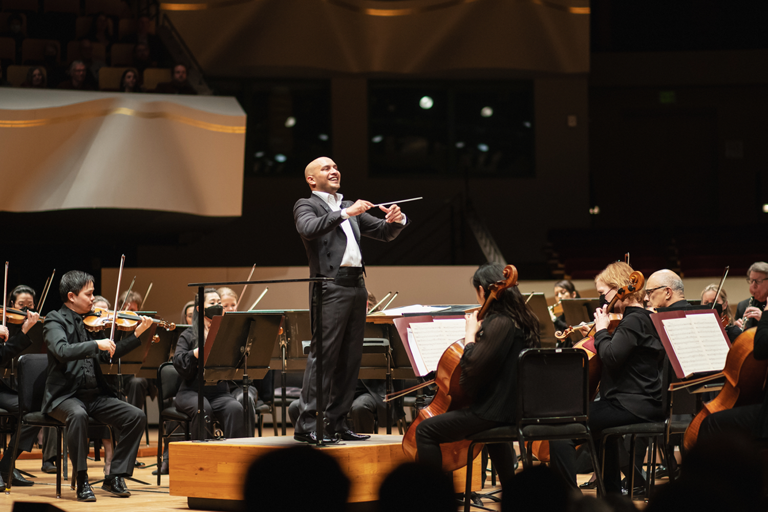 Christopher Dragon stands on a podium at Boettcher Concert Hall, smiling and conducting the orchestra mid-performance, as musicians in formal black attire are playing their instruments around him under warm stage lighting.