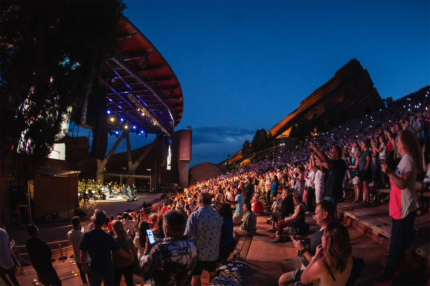 A wide-angle evening shot of a sold-out performance at Red Rocks Amphitheatre in July 2023, featuring Christopher Dragon conducting with Al Green. The audience — thousands of people filling the stone terraced seating — are on their feet, many with arms raised and phones held up. The lit stage is visible on the left, where the orchestra and performers are silhouetted under blue stage lights and a large video screen. Red Rocks' signature towering sandstone monoliths glow amber on the right against a deep blue dusk sky. Photo credit: Amanda Tipton.