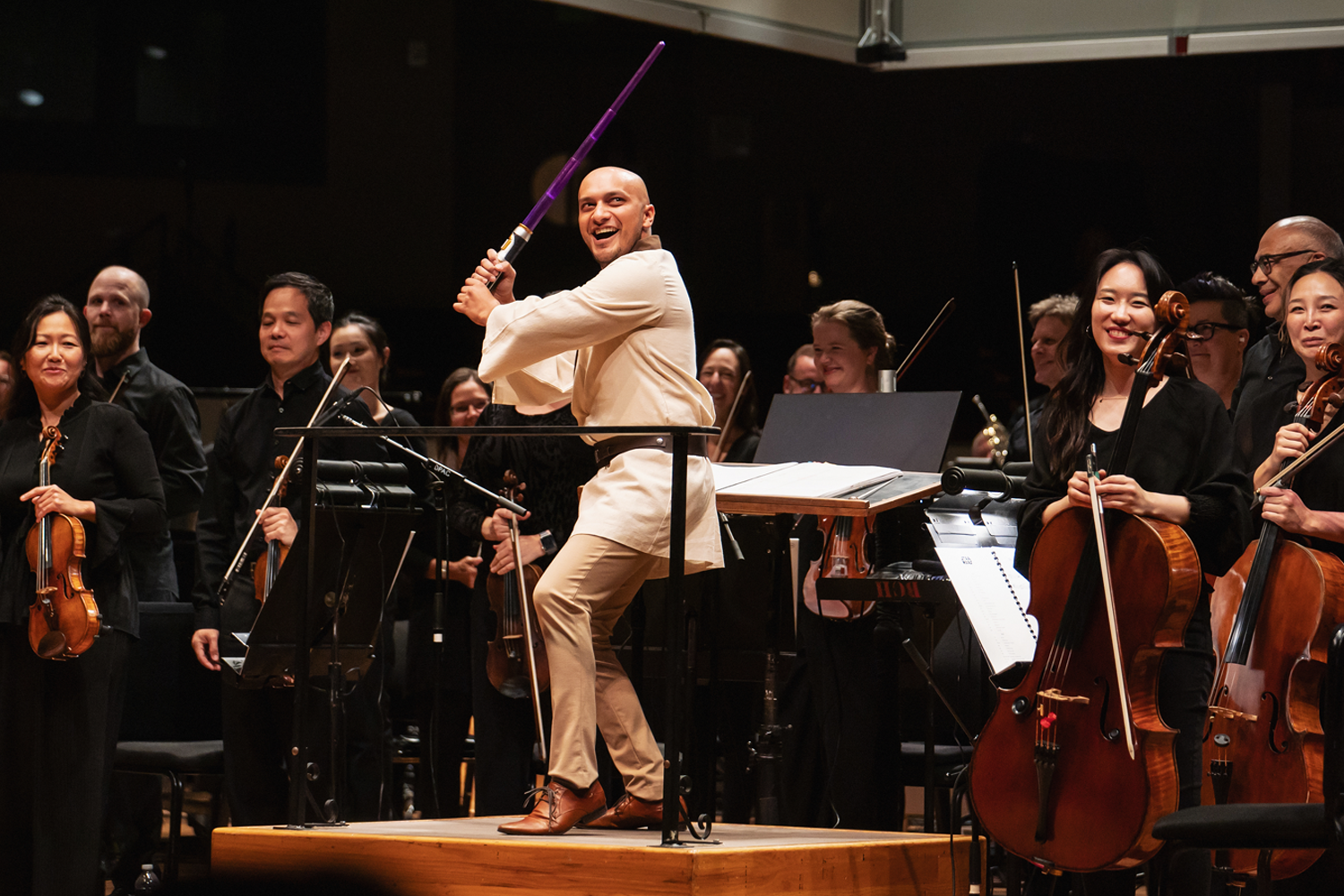 Prior to Star Wars: A New Hope in Concert Christopher Dragon dressed as Luke Skywalker playfully poses on the podium holding a glowing purple lightsaber, while smiling orchestra musicians are standing on stage holding their instruments. Photo Credit Amanda Tipton.