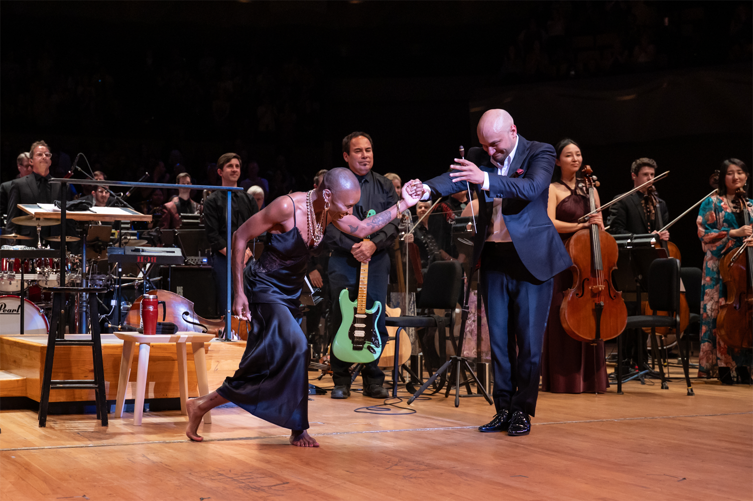 Christopher Dragon holds the hand of Cynthia Erivo as she takes a deep, graceful bow on the stage of Boettcher Concert Hall in May 2025. Erivo, barefoot and wearing a navy satin slip dress with layered necklaces, bends low while Dragon, dressed in a navy suit, leans forward to present her to the audience. A guitarist holding a mint green electric guitar stands behind them, smiling, as orchestra musicians — including cellists — look on approvingly. A Pearl drum kit and other instruments are visible in the background. Photo credit: Amanda Tipton.