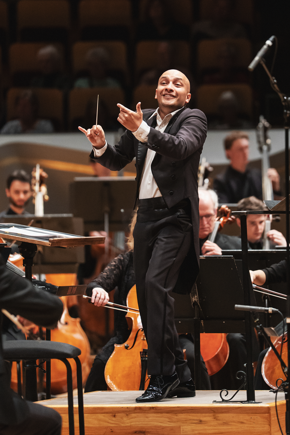 Christopher Dragon conducting with an expansive, open gesture — arms spread forward and smiling broadly toward the orchestra, baton in his right hand. String players and their instruments are visible behind him. Photo Credit Amanda Tipton.