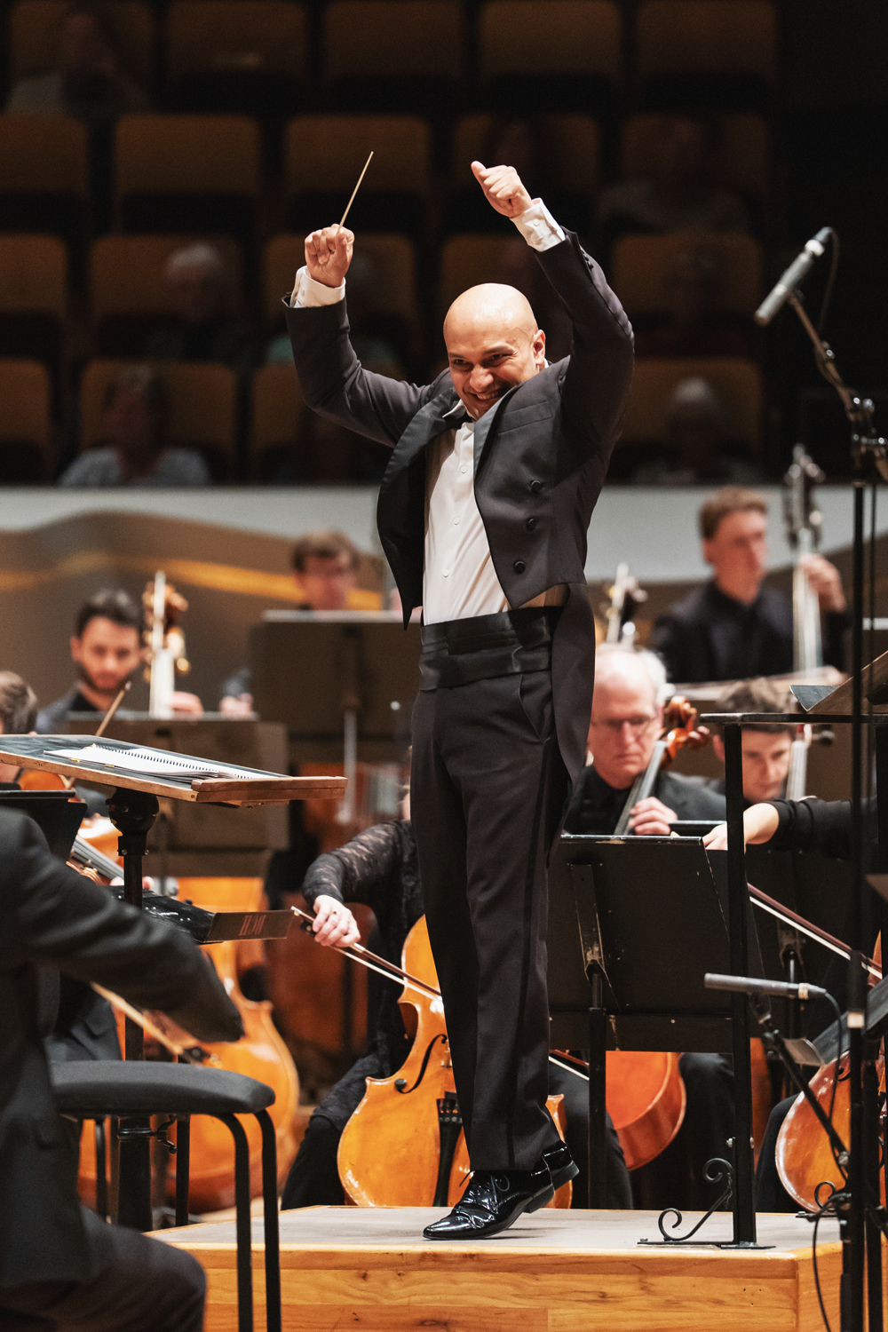 Christopher Dragon at a peak conducting moment, both arms raised triumphantly overhead as if about to jump with baton in hand, body fully extended in an expressive, exuberant pose on the podium. Orchestra musicians are visible in the background. Photo Credit Amanda Tipton.