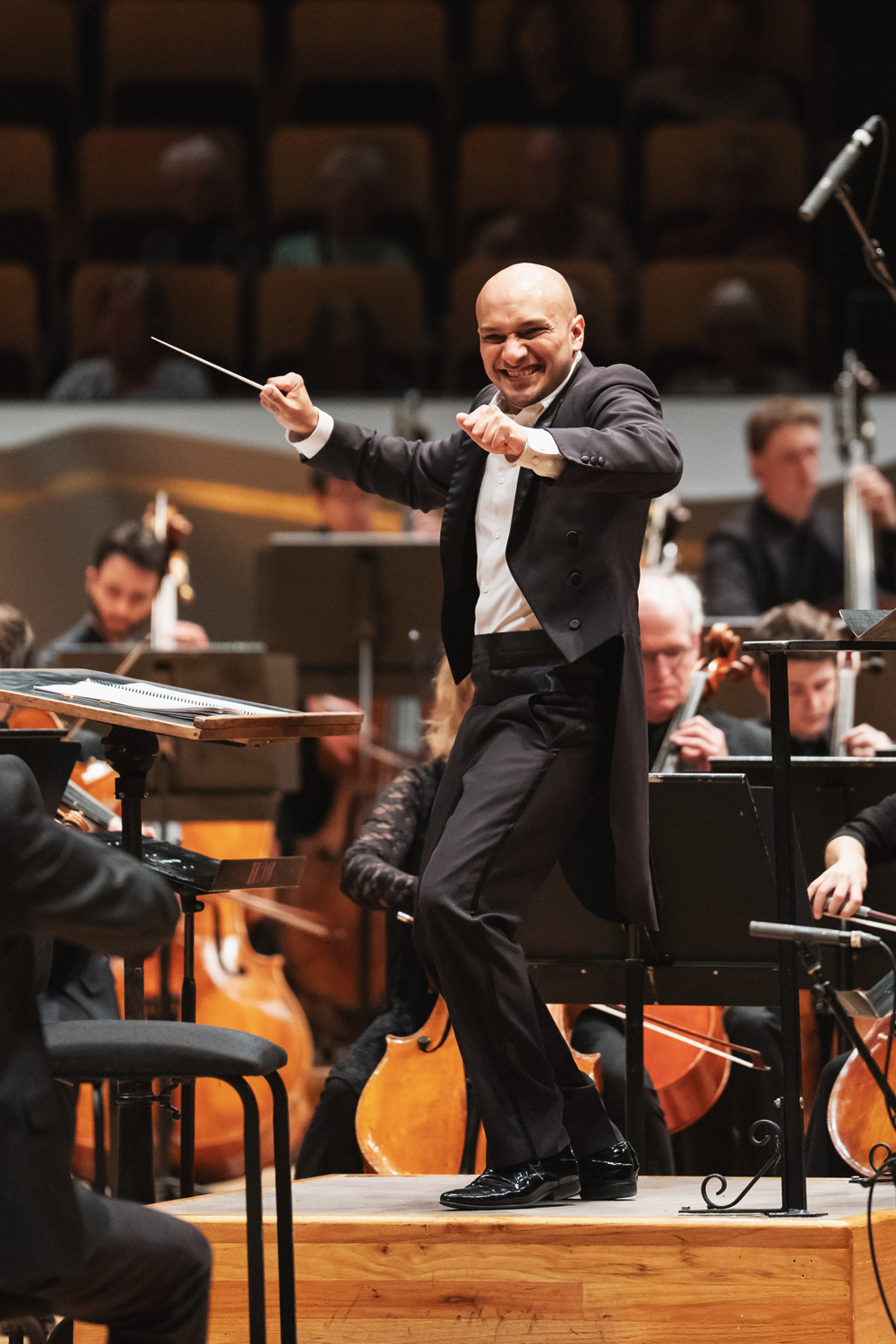 Christopher Dragon conducting with energy and joy. His knees are bent slightly as if he just stomped his foot and his baton arm extended outward and a wide smile on his face. String musicians and the warm wood tones of the concert hall are visible behind him. Photo Credit Amanda Tipton.