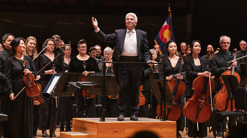 Music Director Peter Oundjian stands with his hand raised atop the podium on stage while facing the audience during applause. The orchestra members are standing behind him and smiling.