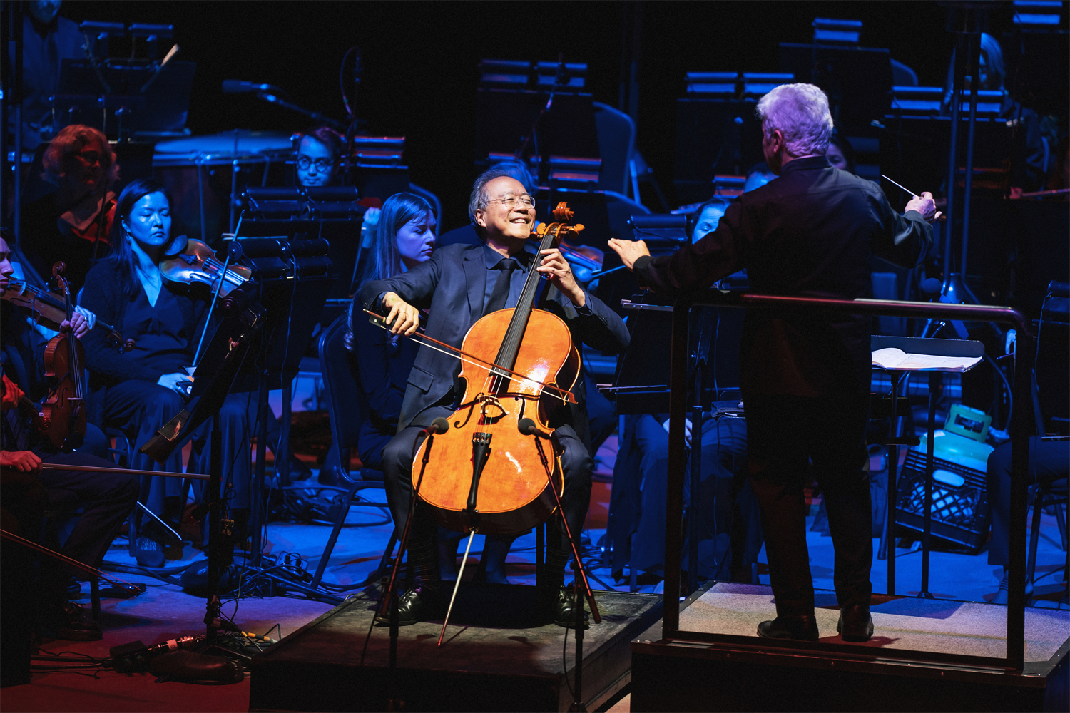 Yo-Yo Ma performs cello at Red Rocks Amphitheatre, bathed in warm amber stage light that makes the rich orange wood of his instrument glow vividly against the cool blue stage lighting surrounding the orchestra. He sits center stage with a wide, radiant smile, deeply engaged in the music. Music Director Peter Oundjian stands with his back to the camera at the right, baton raised toward the orchestra. Two microphones are positioned close to the cello's body to capture its sound in the outdoor venue. Orchestra musicians are arranged in the background, illuminated in moody blue light. Photo credit: Amanda Tipton Photography.