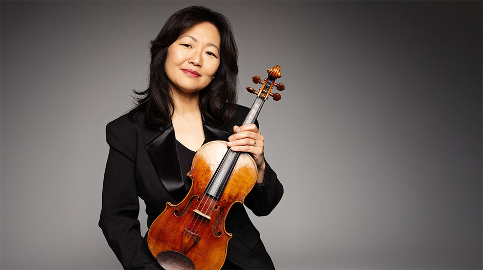 Colorado Symphony Concertmaster Yumi Hwang-Williams poses holding her violin, wearing a black blazer in front of a gray background.