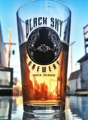 Image of Black Sly Brewery glass, about half full of beer, featuring a city scape and blue skies in the background.