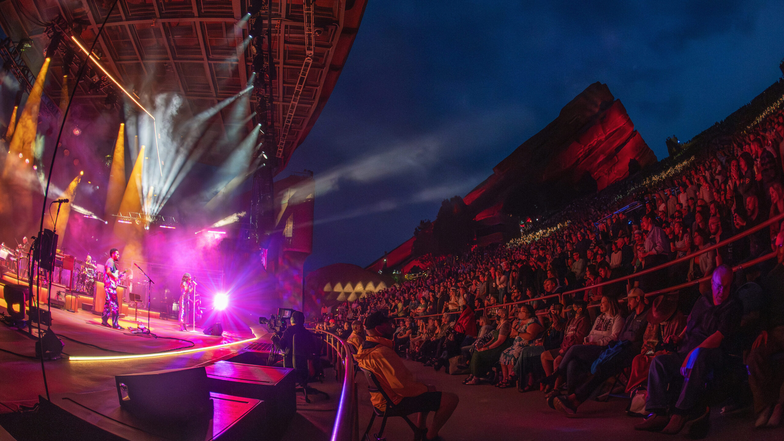 The Colorado Symphony performs with Lake Street Dive at Red Rocks Amphitheatre in Morrison, CO. The sky is dark in the background, and the audience is lit up in red light. The stage features two musicians and a bright pink light.
