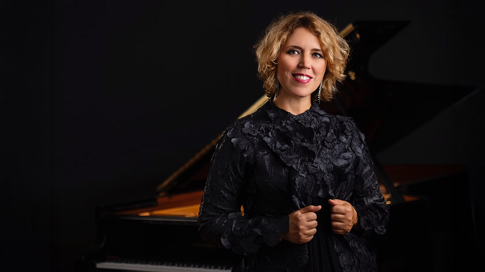 Pianist Gabriela Montero stands in front of a piano while wearing a black lace top.