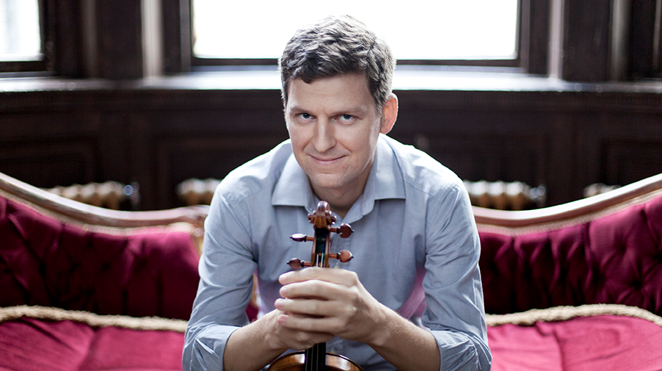 Violinist James Ehnes poses holding his violin wearing a blue shirt while sitting on a red velvet sofa.