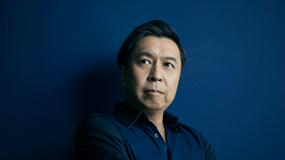 Pianist Makoto Ozone poses wearing a dark blue shirt in front of a dark blue background.