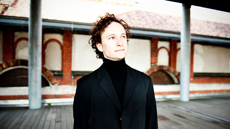 Pianist Martin Helmchen stands in a train station while wearing a black turtleneck and suit jacket.