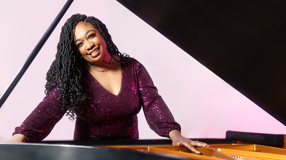 Pianist Michelle Cann stands in behind a piano while wearing sparkly purple dress, in front of a light purple background.