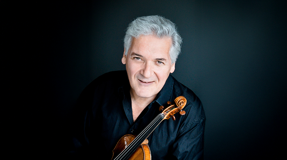Violinist Pinchas Zukerman posts with his violin while wearing a black shirt in front of a black background.