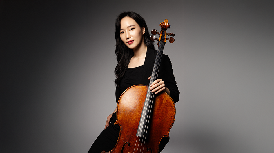 Cellist Seoyoen Min poses with her cello wearing concert black in front of a well lit gray background.