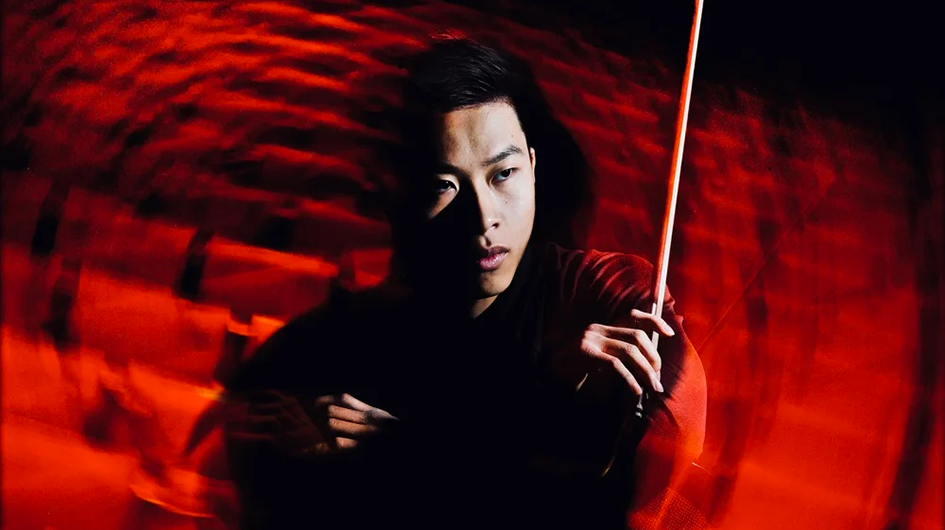 Violinist Kerson Leong poses with his violin bow in front of a blurred bright red auditorium.