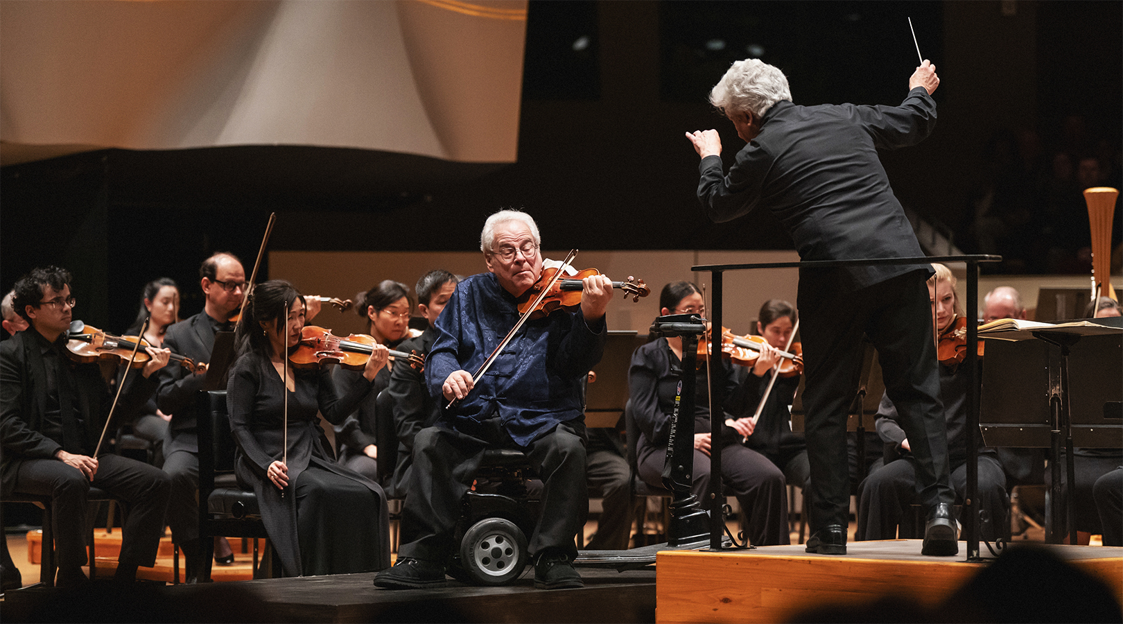 Itzhak Perlman is playing violin on stage at Boettcher Concert Hall. Peter Oundjian is leaning towards Perlman as he conducts. The orchestra is behind them both.