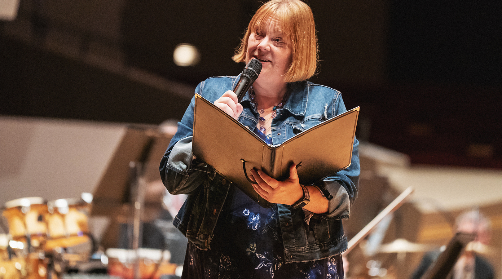History Colorado’s Shaun Boyd is speaking into a microphone onstage at Boettcher Concert Hall. She is holding a microphone to her mouth and a black folder in her other hand. She is wearing a blue jean jacket over a blue floral dress.