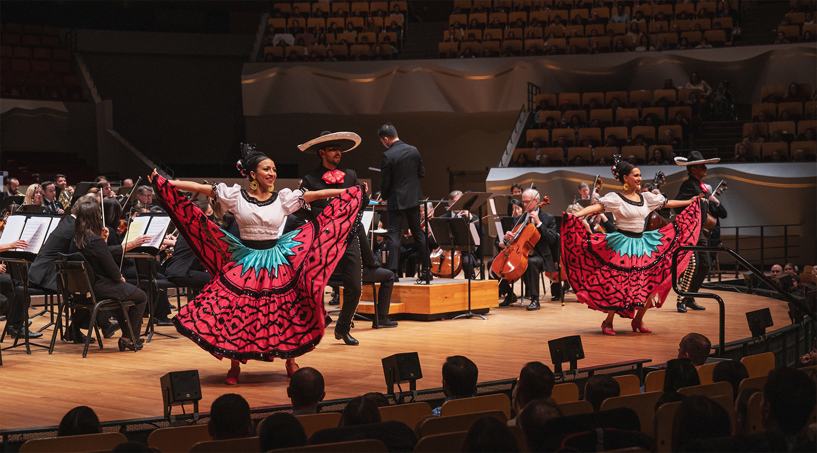 Members of ArtistiCo Dance Company are performing Folklorico dance on stage at Boettcher Concert Hall during the Youth Concert. The women are wearing traditional Jalisco dresses with pinkish-red skirts and ruffled black ribbon. The men are wearing traditional black Charro suits.