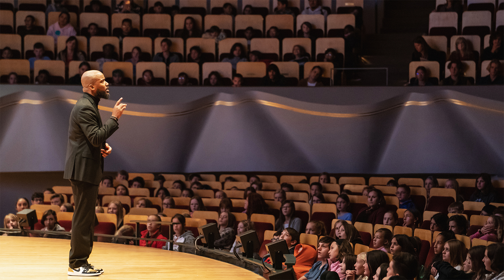 Frankie Le’Troy performs his spoken word poem “Lift Every Voice” while on stage at Boettcher Concert Hall during the Youth Concert. He is a black man wearing a black suit, black turtleneck, and sneakers. His right hand is half raised and the student audience can be seen looking and listening to him.