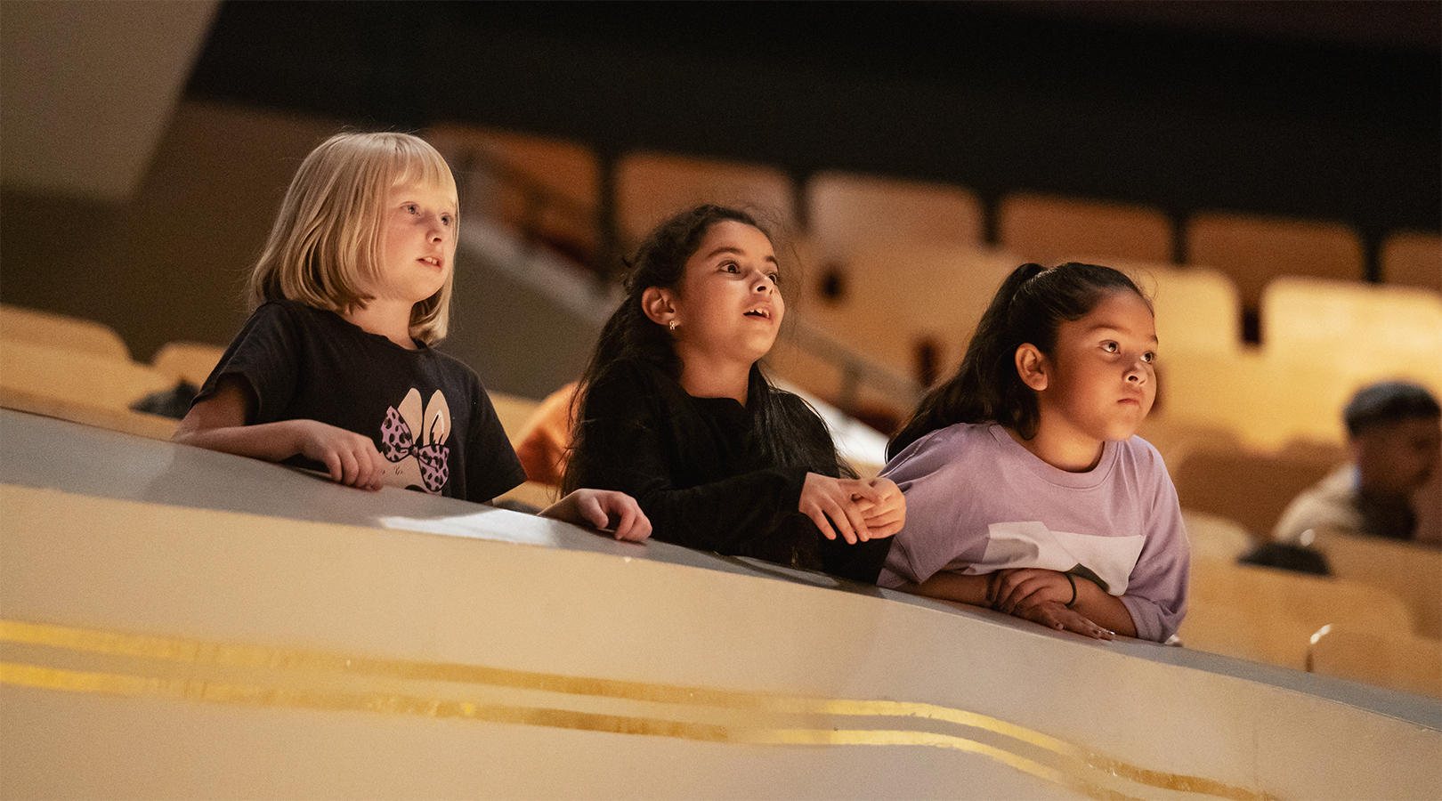 Three students are leaning over one of the balconies at Boettcher Concert Hall during a Youth Concert. They are looking with awe and wonderment.