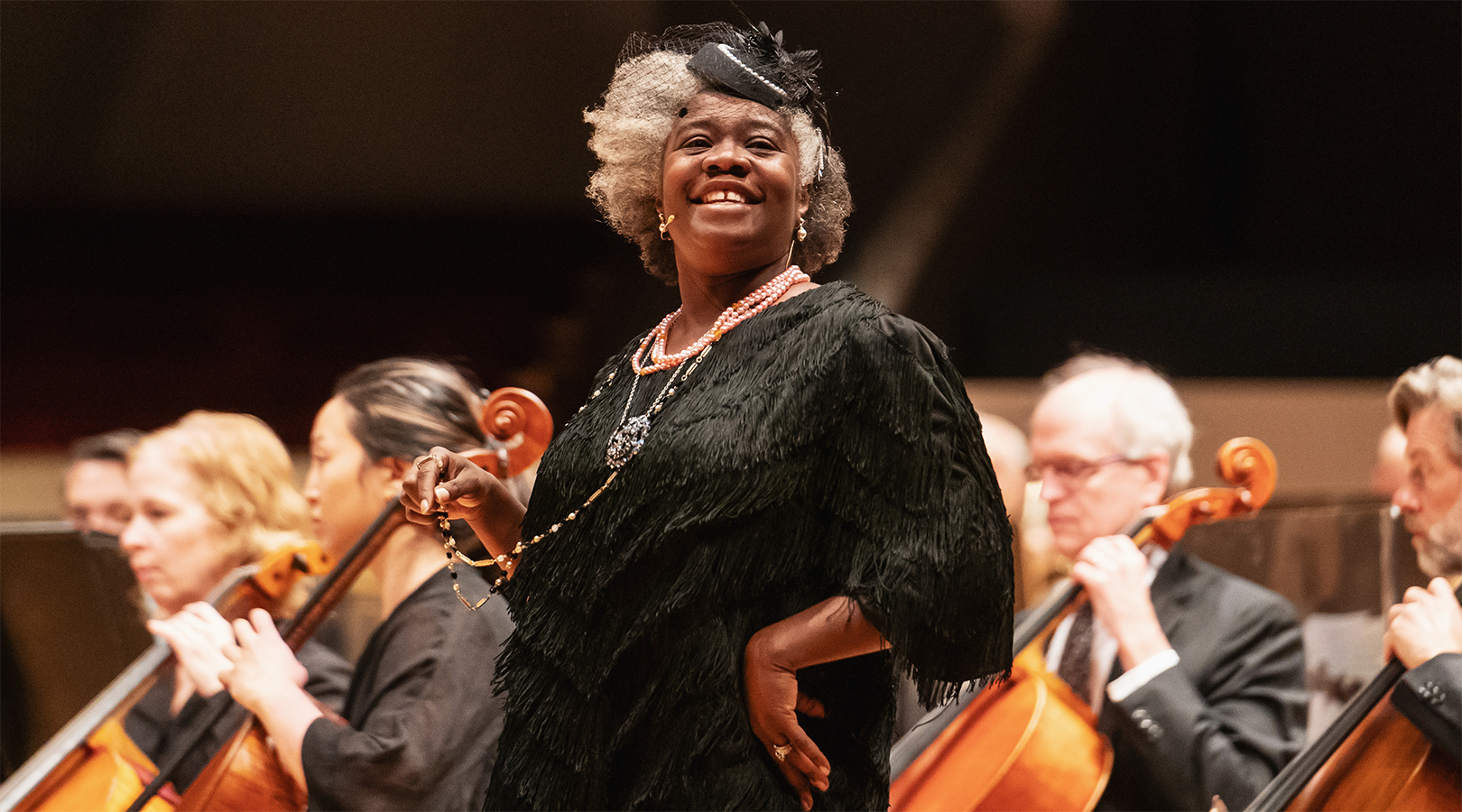 A closeup image of a member of the Black American West Museum and Heritage Center’s Jane Taylor Reenactors Guild performing on stage at Boettcher Concert Hall during the Youth Concert. She is a black woman wearing a black dress and a small black hat. She has short grey hair and a bright smile.