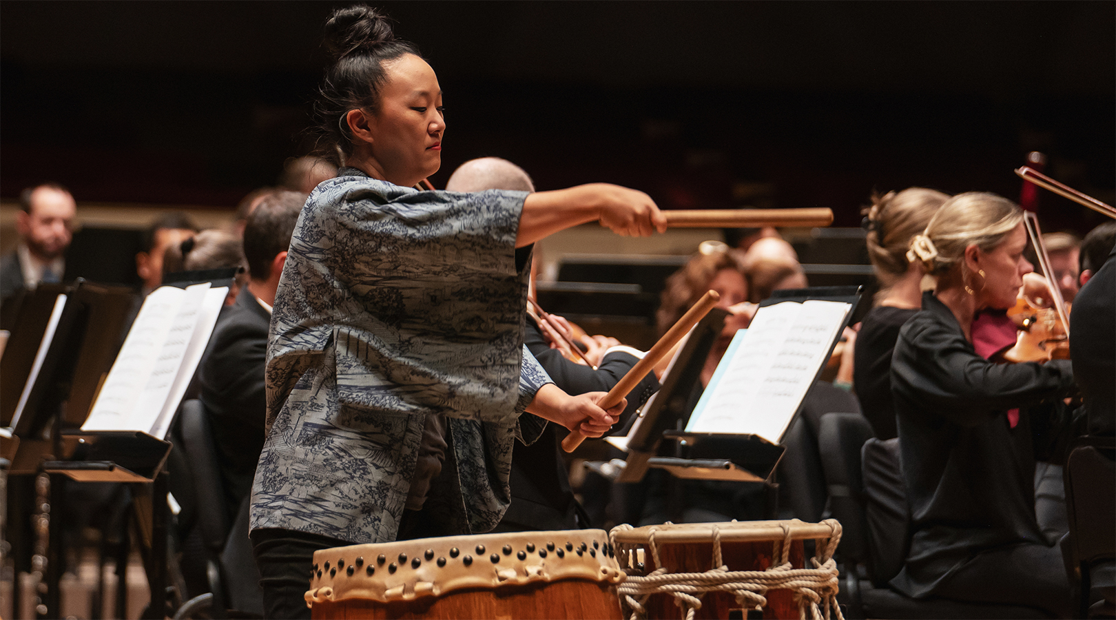 Courtney Ozaki-Durgin from the Japanese Arts Network performing on stage at Boettcher Concert Hall during the Youth Concert. She has her hair pulled back in a high bun, is wearing a blue top, and is performing the traditional  Taiko drum.