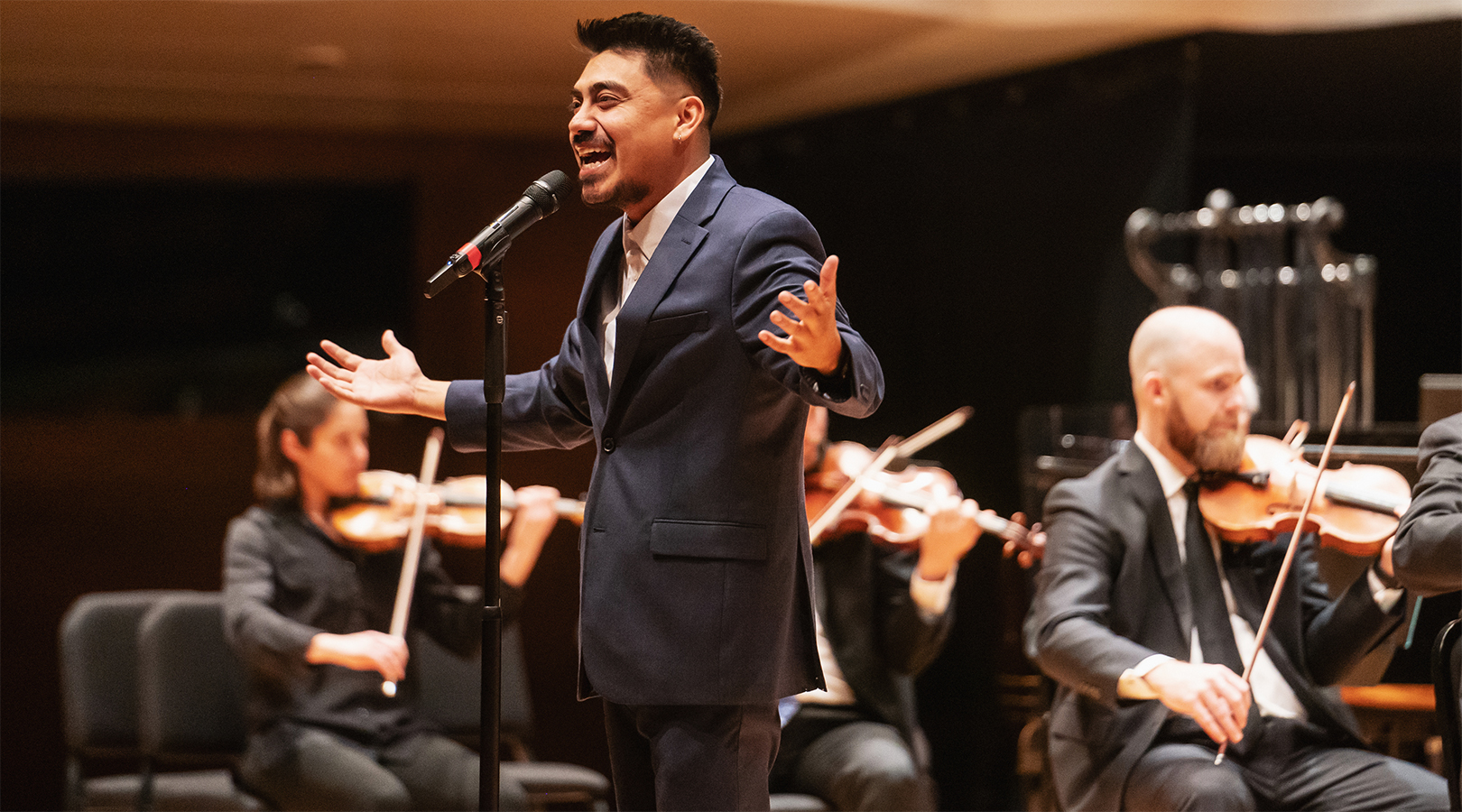 Spoken word poet, José “Jozer” Guerrero is speaking into a standing microphone while on stage at Boettcher Concert Hall during a Youth Concert. He is wearing a blue suit and his hands are outstretched towards the student audience.