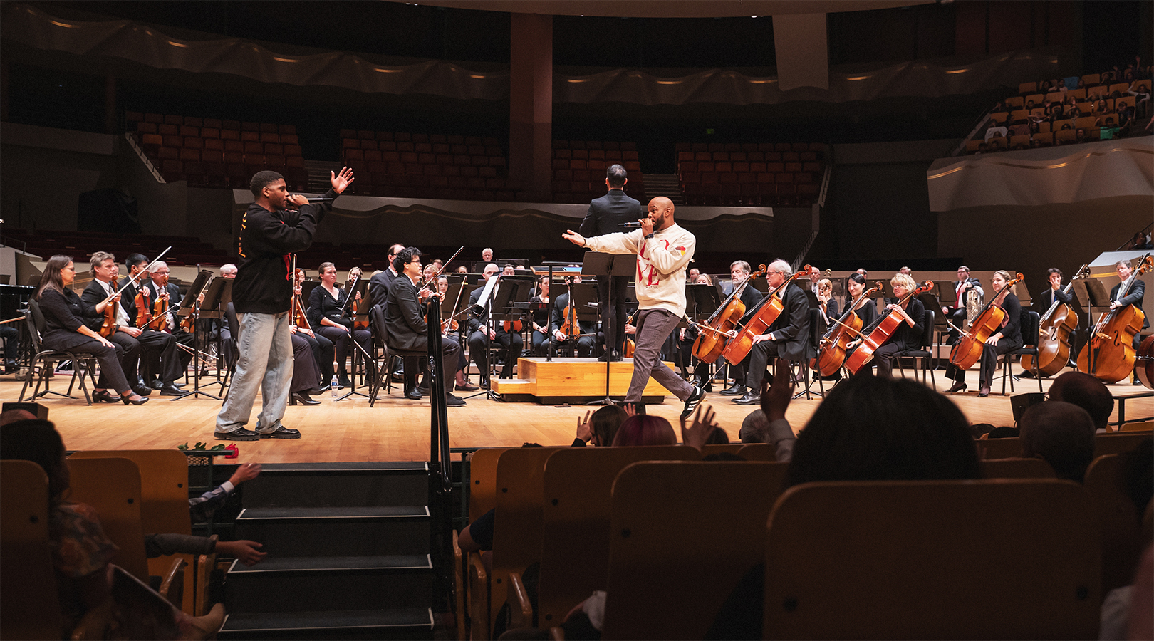 Chinelo “Nelo” Tyler and Wayne Watts performing on stage at Boettcher Concert Hall during the Youth Concert. Tyler is wearing a black sweatshirt and blue jeans while holding a microphone to his mouth and rapping. Watts is wearing a white sweatshirt with the word "Love" in red across the chest, also holding a mic to his mouth and rapping. They are both facing each other with one arm extending outward. The orchestra is in the background performing.