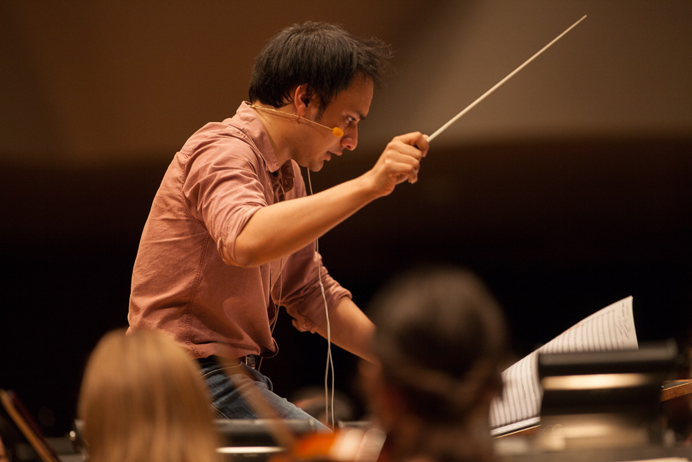 Christopher Dragon conducting during a rehearsal in January 2016, captured in a close-up side profile shot. He leans forward with clear intensity and concentration, baton held aloft in his right hand, wearing a casual pink button-down shirt and jeans with a headset microphone. Orchestra musicians and a music stand with sheet music are softly blurred in the foreground.