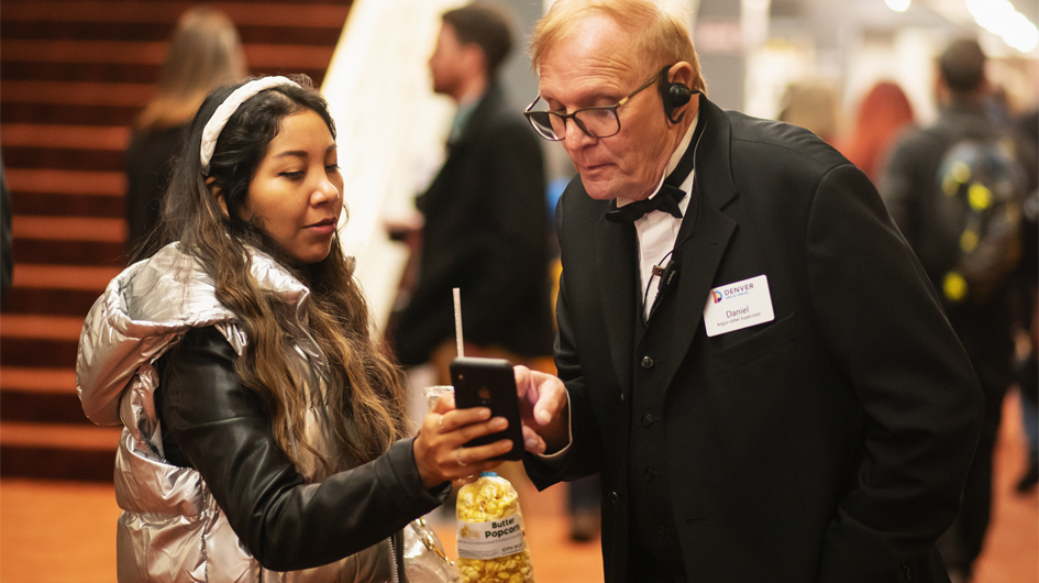 An usher holding programs is pointing across the house in Boettcher Concert Hall and helping a parent holding a child find their seats.