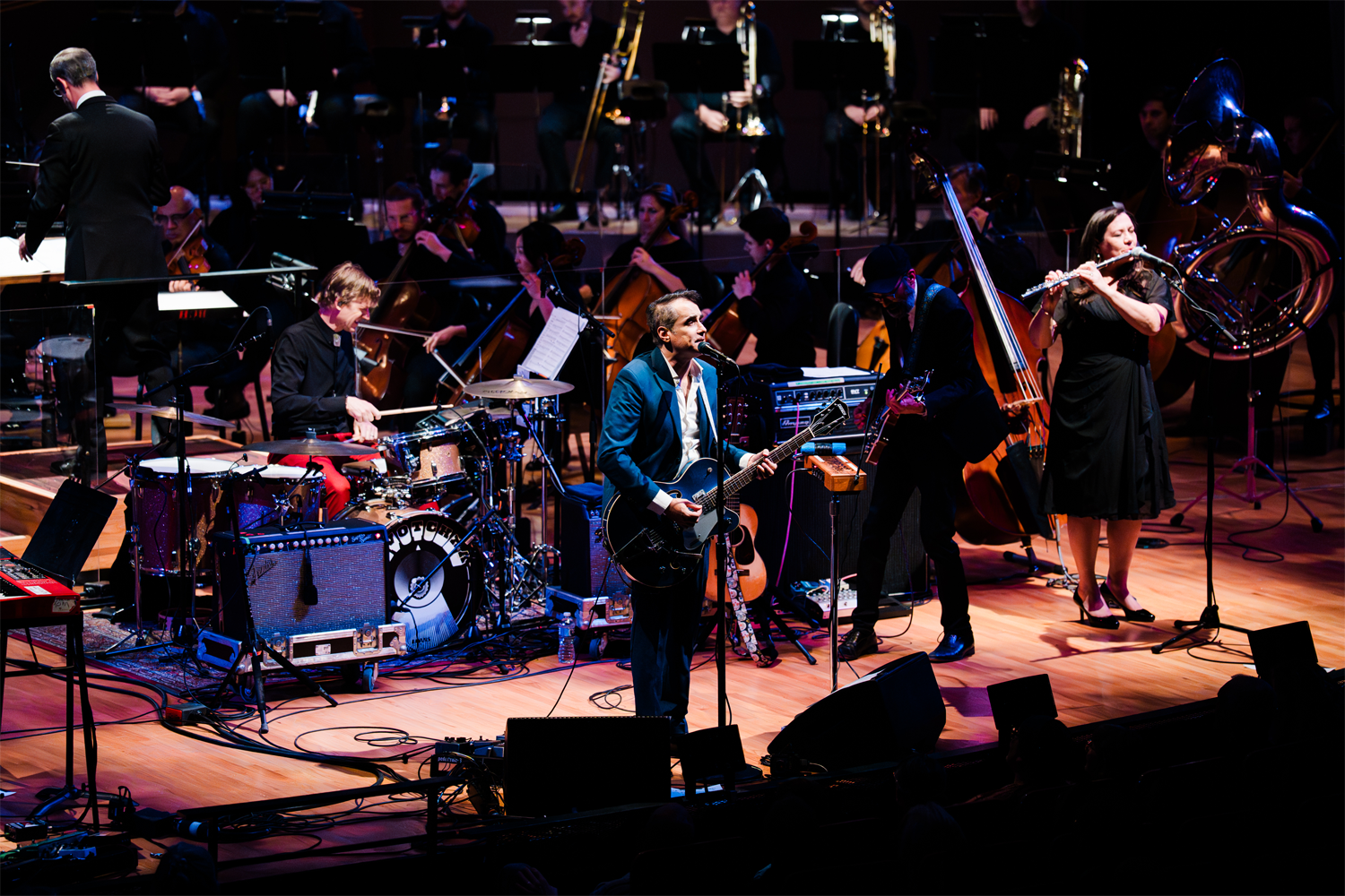 DeVotchKa performs with the Colorado Symphony in November 2025 on the stage of Boettcher Concert Hall. In the foreground, the band's lead singer Nick Urata stands at a microphone playing a dark electric guitar, wearing a teal blazer. To his left, the drummer Shawn King plays a kit with a decorative bass drum head, and to his right, Tom Hagerman plays bass guitar and Jeanie Schroder plays flute while a large sousaphone gleams in the stage light. Behind the band, the full symphony orchestra is arranged across the stage, with string players, brass, and a conductor visible at the far left. The warm wood floor of the stage is crisscrossed with cables and monitor speakers, capturing the organized energy of a genre-blending live performance.