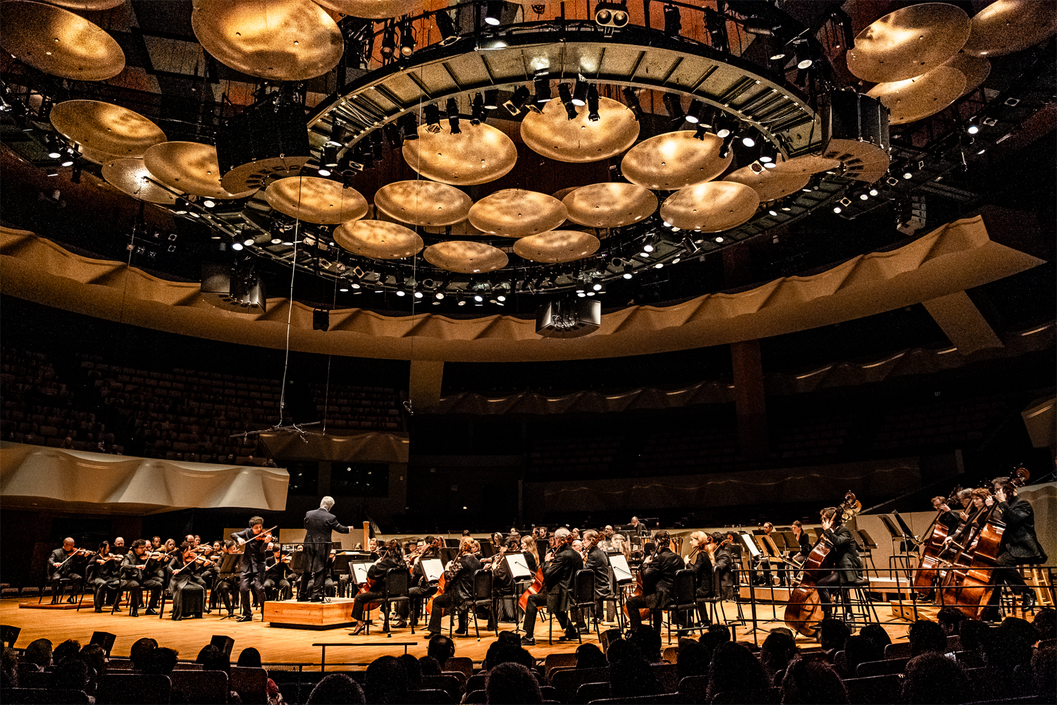 Peter Oundjian conducts the Colorado Symphony as Augustin Hadelich performs Tchaikovsky's Violin Concerto at Boettcher Concert Hall in May 2025. The photo is taken from an elevated side angle, looking down toward the stage where the full orchestra is arranged around the conductor's podium. Boettcher's iconic ceiling installation — concentric rings of large, golden bowl-shaped acoustic reflectors interspersed with stage lighting — dominates the upper portion of the frame in rich amber tones. The surrounding hall walls feature the venue's signature sculptural, faceted paneling. A small audience is visible in the darkened foreground seating.