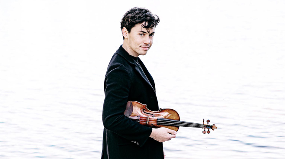 A relaxed, atmospheric promotional portrait of violinist Benjamin Beilman, photographed outdoors in front of a calm, pale body of water that blends softly into a hazy sky. He stands in a three-quarter profile, holding his violin loosely at his side, and turns his head to look back toward the camera with a quiet, assured expression. He wears a black velvet blazer and has dark, loosely curled hair. The muted, ethereal tones of the background give the image a dreamy, understated elegance.