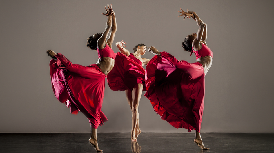 A stunning performance photo of three dancers from Cleo Parker Robinson Dance, captured mid-movement against a warm neutral gray backdrop. The two outer dancers mirror each other in sweeping arabesque-like poses, arms reaching skyward with fingers splayed, while their voluminous red skirts billow outward in motion. The center dancer, in a shorter red costume, balances on one leg in a more contained pose with arms floating gently. All three are barefoot. The rich crimson of the costumes against the soft gray background creates a striking, painterly image full of motion, symmetry, and grace. Photo credit: Jerry Metellus.