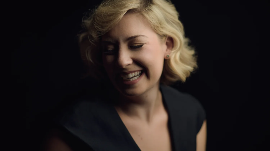 A warm, intimate promotional portrait of soprano Diana Newman, captured mid-smile with her eyes cast slightly downward, conveying a natural, joyful ease. She wears a dark navy or black sleeveless top and a delicate necklace, with short, softly waved blonde hair framing her face. The image is lit with a single soft light source that illuminates her face and shoulders against a near-black background, giving the portrait a classic, chiaroscuro quality that feels both elegant and approachable.