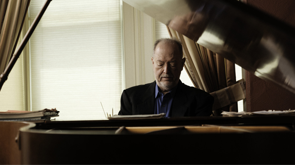 A moody, intimate portrait of pianist Garrick Ohlsson seated at a grand piano, shot from the perspective of across the open piano lid. He looks downward in calm concentration, wearing a black blazer over a blue shirt. Sheet music and papers are stacked on both sides of the piano. Soft natural light filters through a window with white blinds behind him, and the blurred piano lid fills the upper right of the frame, adding depth and an artistic, immersive quality to the image.