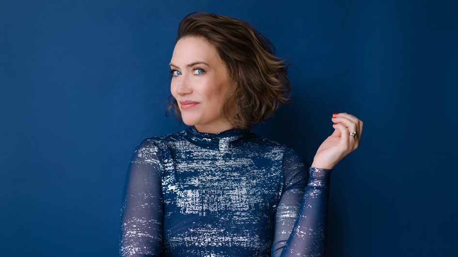 A vibrant, fashion-forward promotional portrait of soprano Heidi Stober, posed against a rich cobalt blue wall that complements her long-sleeved, sheer blue sequined dress. She leans slightly back with one hand raised gracefully behind her shoulder, red-painted nails catching the light, and looks off to the side with bright blue eyes and a relaxed, confident smile. Her short, tousled brunette hair adds a sense of movement to the image. The tonal harmony of the blue dress against the blue wall creates a striking, cohesive composition with a modern, editorial feel.