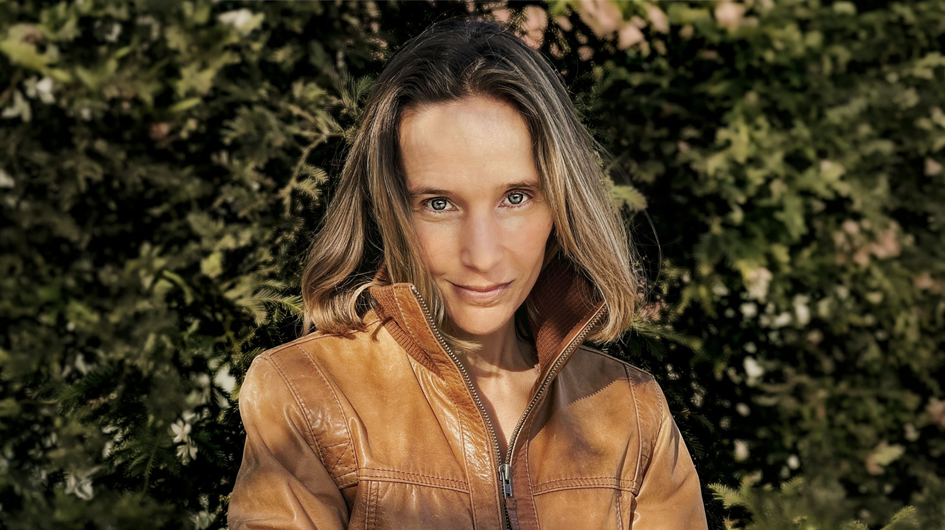 A natural, warmly lit outdoor portrait of pianist Hélène Grimaud, photographed against a full backdrop of dense green foliage. She faces the camera directly with a calm, steady gaze and a subtle, understated expression. Her shoulder-length, light brown hair catches the sunlight, and she wears a warm tan leather zip-up jacket. The dappled natural light and organic background give the portrait an earthy, unguarded quality that sets it apart from more formal classical musician photography.
