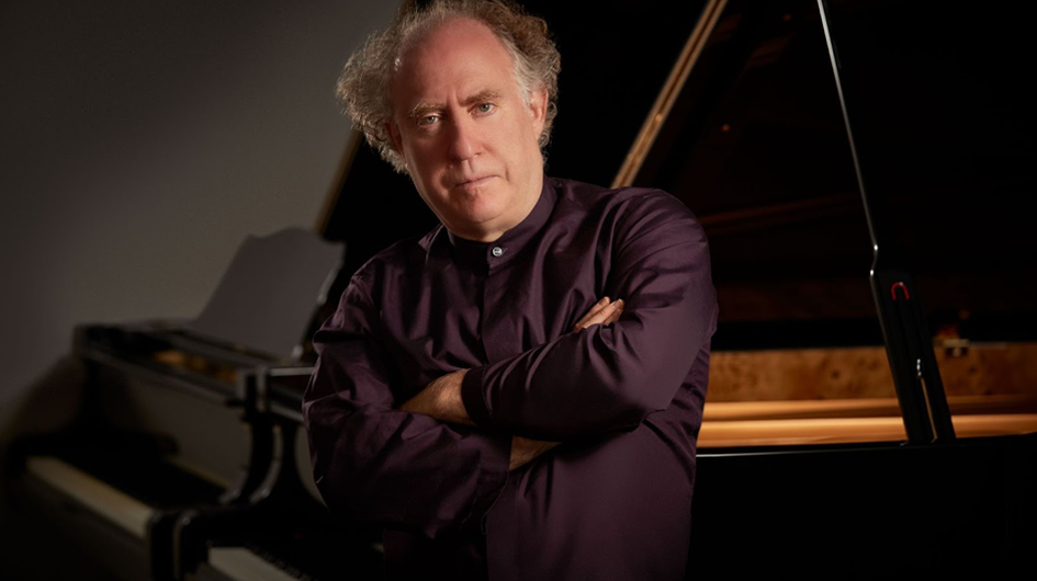 A confident, moody promotional portrait of pianist Jeffrey Kahane, seated and leaning back against an open grand piano with arms folded across his chest. He gazes directly into the camera with a composed, steady expression. He wears a deep plum/burgundy mandarin-collar shirt. His curly salt-and-pepper hair and the warm wood interior of the piano's open lid are illuminated against a dark gray background, giving the image a rich, dramatic quality that balances authority with artistic warmth.