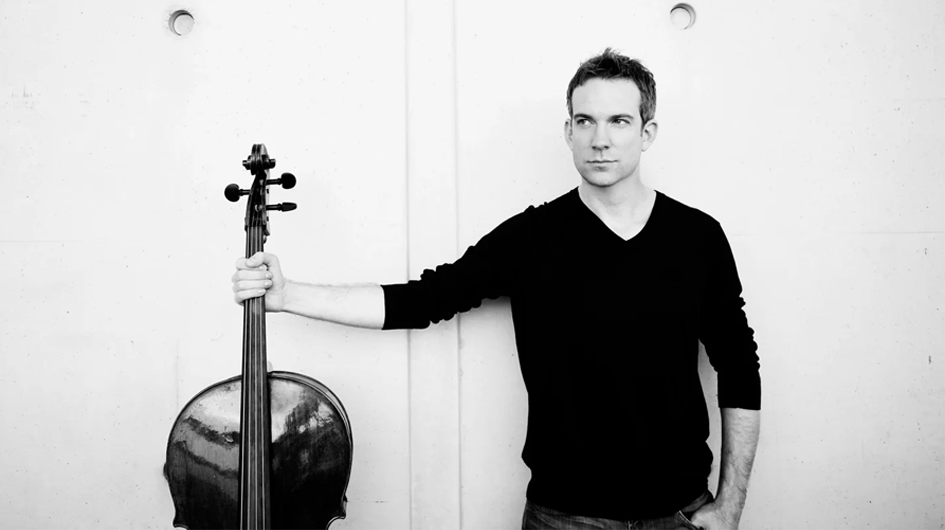 A striking black and white promotional portrait of cellist Johannes Moser. He stands casually against a pale paneled wall, one arm extended to grip the neck of his cello — which rests upright beside him — while his other hand is tucked into his jeans pocket. He wears a fitted black V-neck sweater and gazes off to the side with a calm, composed expression. The minimalist composition and high-contrast monochrome treatment give the image a timeless, editorial quality. Photo credit: Sarah Wijzenbeek.