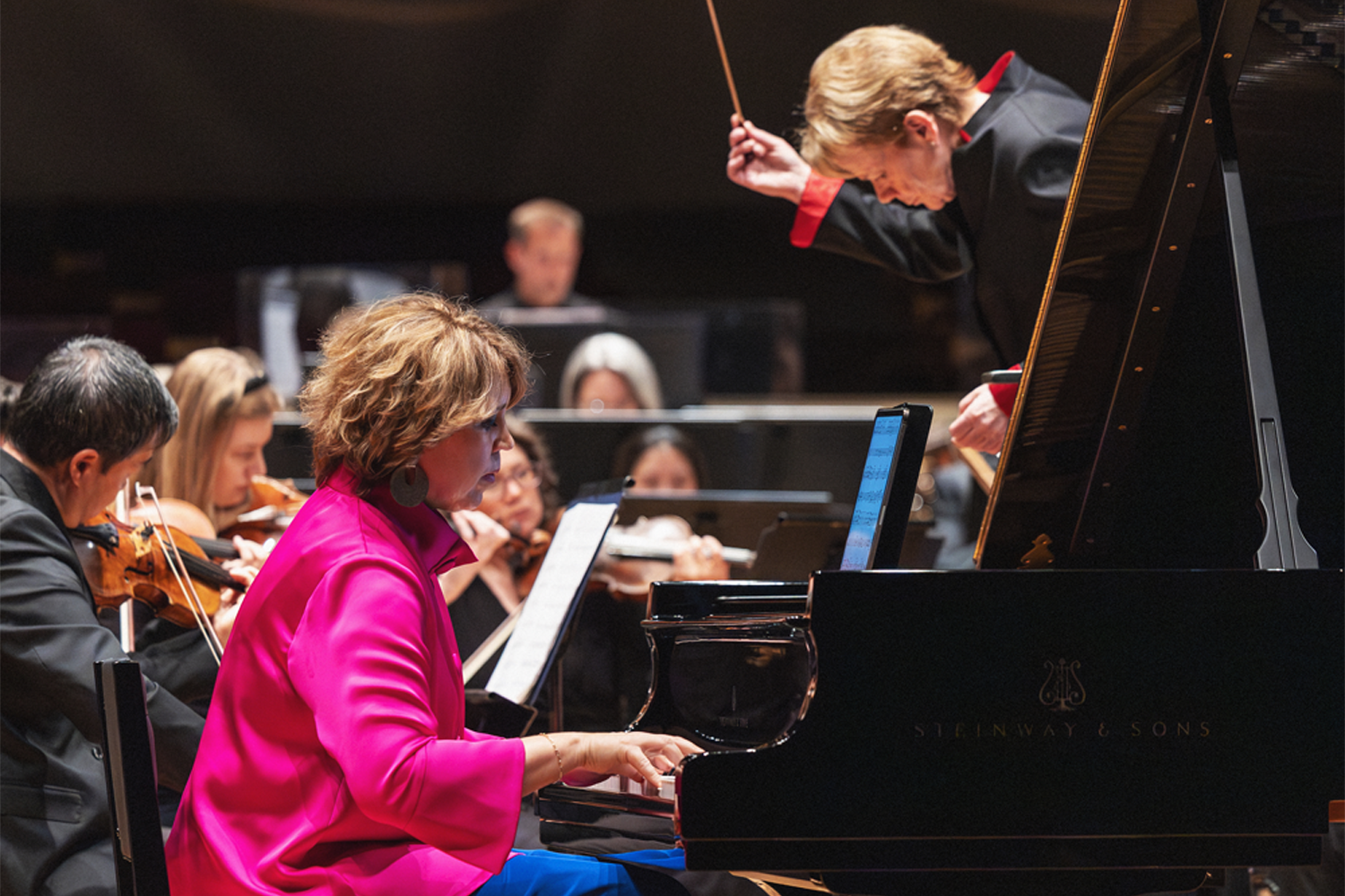 A dynamic, close-up performance shot of pianist and composer Gabriela Montero at the keyboard of a Steinway & Sons grand piano, dressed in a vivid hot pink satin top and blue trousers, her expression one of intense focus as she reads from the score. Above her, conductor Marin Alsop leans dramatically over the piano lid, baton raised mid-gesture, wearing a black jacket with red lining. The physical closeness of the two artists creates a compelling sense of musical dialogue. String players and other orchestra musicians are visible in soft focus in the background. Photo credit: Amanda Tipton Photography.