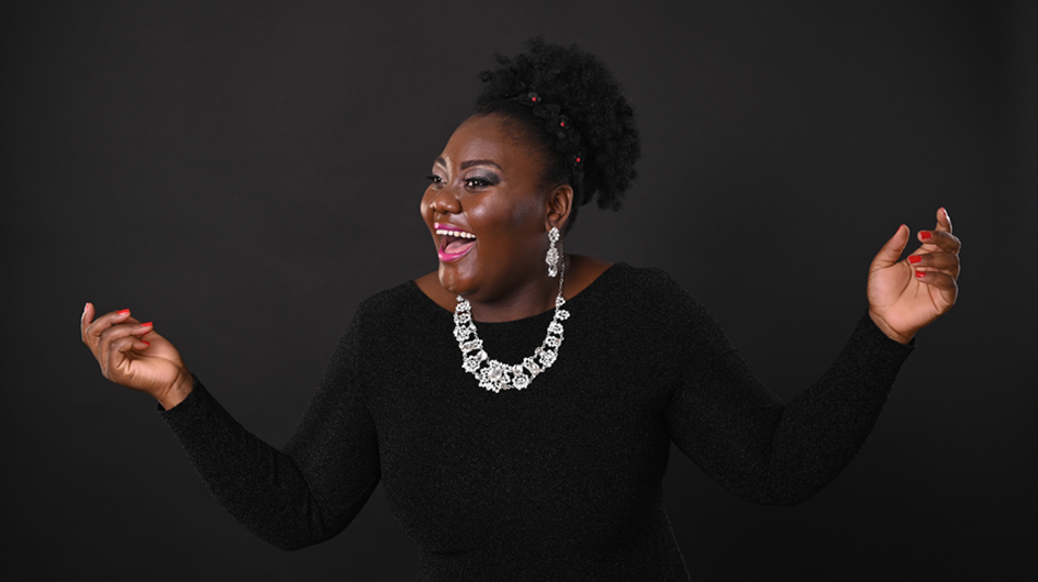 A radiant, expressive promotional portrait of soprano Masabane Cecilia Rangwanasha, captured mid-laugh with both arms spread wide and palms open, conveying warmth and exuberance. She wears a black long-sleeve top, a statement crystal floral necklace, matching chandelier earrings, and red nail polish, with her natural hair swept up and adorned with small red pins. Her bright pink lipstick and joyful expression give the image an infectious, celebratory energy against the deep charcoal black background. Photo credit: Vera Elma Vacek.
