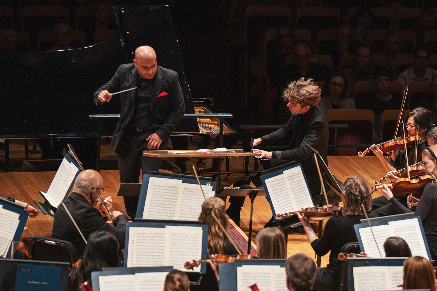 An immersive, overhead-angle performance shot of Resident Conductor Christopher Dragon and pianist Albert Cano Smit in an intense moment of musical dialogue on the Boettcher Concert Hall stage. Dragon leans forward from the left with baton raised, wearing a black suit with a red pocket square, his focus directed toward Smit, who sits at the Steinway grand piano with tousled hair, deeply absorbed at the keys. The foreground is filled with the backs of string musicians' heads and a sea of blue score folders on music stands, drawing the eye inward toward the two soloists at the center of the action. Audience members are faintly visible in the darkened seating tiers behind the piano. Photo credit: Amanda Tipton Photography.