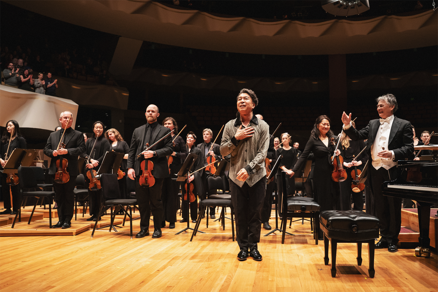 Pianist Makoto Ozone stands at center stage at Boettcher Concert Hall, hand pressed to his heart and face tilted upward with a joyful, grateful smile as he acknowledges the audience's applause following a performance. Conductor Jun Märkl stands to his right in white tie formal attire, one arm raised toward Ozone in a gesture of presentation and celebration. The orchestra musicians — many holding their violins and bows at their sides — are arranged behind them, several smiling and joining in the applause. A grand piano is partially visible at the right edge of the frame. Audience members can be seen applauding from the upper tier seating in the background. Photo credit: Amanda Tipton Photography.