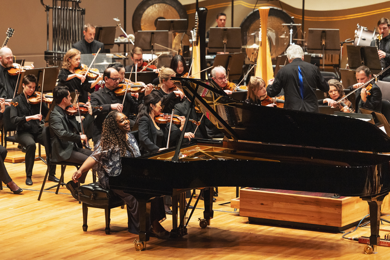 Pianist Michelle Cann is captured in a joyful, candid moment at the Steinway grand piano on the Boettcher Concert Hall stage, turned sideways in her seat with a wide, beaming smile — perhaps sharing a spontaneous moment of delight with the audience or orchestra. She wears a floral patterned dress with long curly hair. Music Director Peter Oundjian stands with his back to the camera at the right, conducting the surrounding orchestra, his blue-accented jacket visible. The full orchestra fills the background — strings, harp, percussion including large gongs, and other sections arranged across the warm wood-toned stage. Photo credit: Amanda Tipton Photography.