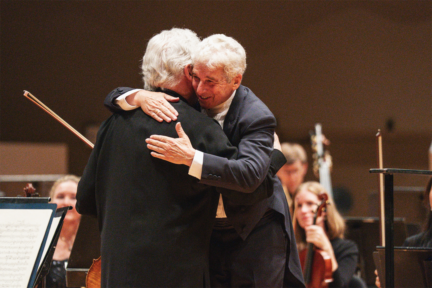 A candid, heartfelt post-performance moment as Pinchas Zukerman and Music Director Peter Oundjian embrace on the Boettcher Concert Hall stage. Oundjian, facing the camera in a navy suit with a broad smile, wraps both arms around Zukerman, whose back is to the camera and who still holds his violin bow in one hand. The genuine warmth between the two longtime colleagues is palpable. In the softly blurred background, smiling orchestra musicians — including a violinist looking on with delight — are visible against the warm brown tones of the concert hall walls. Photo Credit: Amanda Tipton Photography